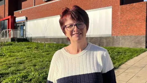 A woman in a navy blue and white knitted jumper smiling. She has short red hair and glasses. She stands on the pavement outside of a building.