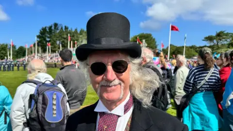 Geoff Robinson a man with long white hair, he wears a top hat and sunglasses and smiles on the field next to Tynwald hill.