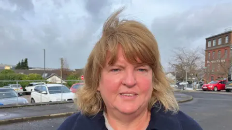 Shows a woman with fair hair and black coat standing in front of cars. 