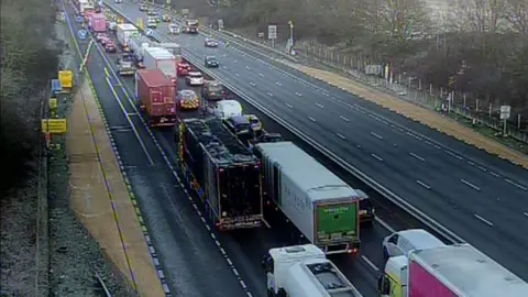 A queue of traffic on one side of the M25, seen from a camera above. There are a large number of lorries and a few cars. The other side of the carriageway is mostly empty, and there are trees growing on either side of the motorway. It looks chilly.