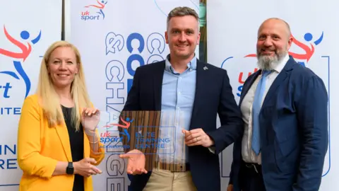 University of Wolverhampton A woman with long blonde hair on the left is wearing a yellow jacket and standing next to a man in the middle with a shirt and dark blue jacket. A man with a grey beard on the right is wearing a shirt, tie and blue jacket.