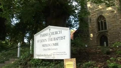 A white sign in front of a small church reading 'parish church of St John the Baptist, Sedlescombe, circa 13th century'.