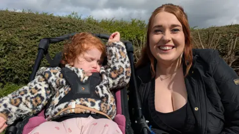 A little girl sitting in a pram. Her mother is smiling at the camera next to her. 