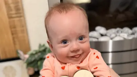 Helen Bennett A baby in a pink babygrow with red hearts on it. She is smiling.