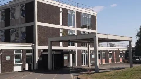 Outside North Devon District Hospital in Barnstaple. The bricks are dark brown. A large white concrete canopy is up above the main entrance.