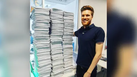 ESO_Danny A young man in a navy blue polo shirt stands next to three huge stacks of A4 paper. They sit on a desk just below the height of his hip and are so tall that they almost reach the top of his head.