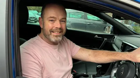 A man in a pink top sitting in a parked car who is smiling at the camera. One hand is on the steering wheel. There are other parked cars in the background.