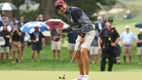 Reuters Whaley stands on a putting green during a rainy outdoor golf event. He is bent slightly forward, holding a putter in one hand while making a clenched-fist gesture with the other hand. He is wearing a maroon cap, a grey and dark navy waterproof jacket, light-colored shorts, and white athletic golf shoes with red accents.