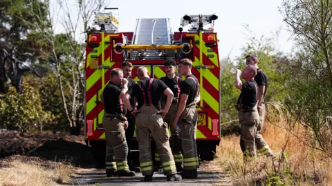 Jacob King/PA Wire Fire service crews gather around a fire engine at the site of the fire. One firefighter is drinking water from a bottle.