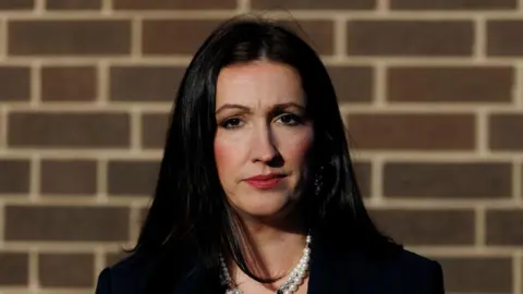 PA Media Little-Pengelly has long dark hair and is wearing make up as she stands against a brick wall. She has a black jacket on and a pearl necklace.