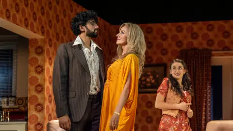 Ellie Kurtz Omar Malik and Tamzin Outhwaite in 1970s costume look at each other on a theatre set depicting a living room. Lauren Patel watches on behind them.