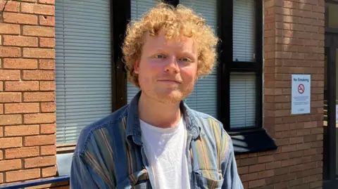 Zac Sherratt/BBC A man with curly ginger hair and ginger stubble standing next to a blue railing.