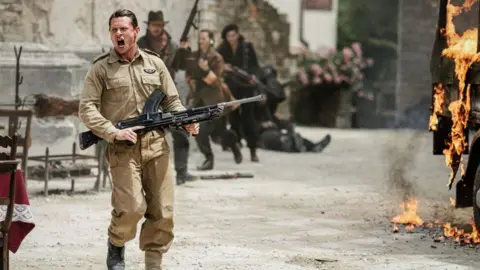 Banijay Rights Jack O'Connell, playing Blair Mayne in Rogue Heroes. He is wearing a khaki military uniform and holding a black rifle. He is shouting as a lorry burns to the right-hand side of the screen.