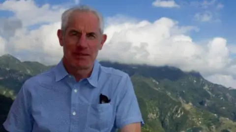 Family photo Tudor, with grey hair in a blue short sleeve shirt. He's standing in front of a mountain range and blue sky and clouds are seen in the background. 