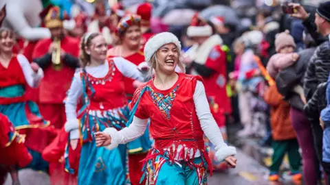 Manchester City Council A woman in a red top and blue skirt grins as she dances and leads a group of other women in white, red and blue outfits at the parade. Spectators line the street, watching and taking pictures.