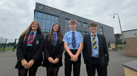 BBC/Jim Scott Four school pupils, wearing a mix of coloured school ties, stand in front of their main school building. In the background is the school's crest and name, High Tunstall College of Science.