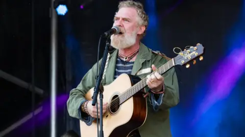 Glen Hansard sings into a microphone while holding an acoustic guitar. He has a white beard and grey short hair. He is wearing a green jacket and a navy and blue striped tshirt.