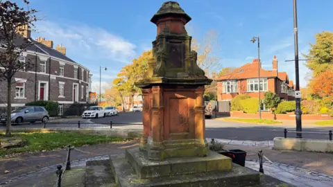 North Tyneside Council The Boer War Memorial is orange and grey. Columns of descending sizes are stacked on top of each other on a plinth. It is located in a square surrounded by semi-detached residential houses.