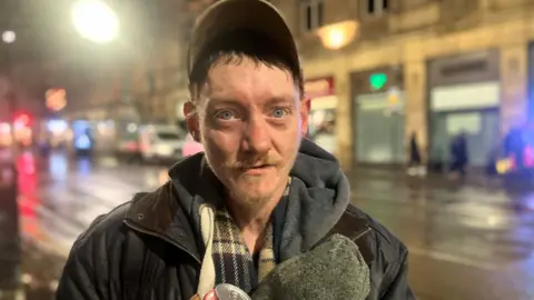 Photograph of Jordan Winstanley (43) from Higher Openshaw in East Manchester. He is pictured on Cross Street after receiving food and warm clothing from a local charity. 