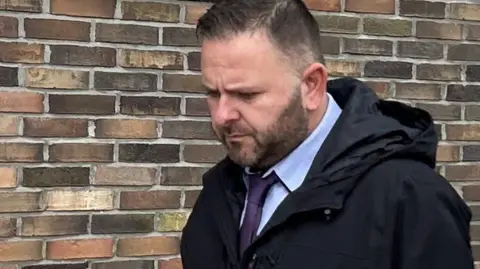 Paul Higgins, frowns and looks toward the floor. He is outside, wearing a coat and dark tie. There is a brick wall behind him