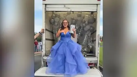 Family photograph Jessica, with long dark hair and black glasses, wears a purple prom dress as she stands on a tail lift on the back of a removals van. In one hand she is holding a white handbag.