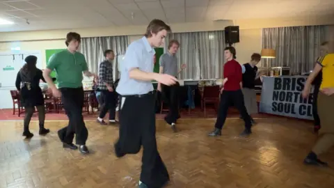 People wearing wide legged trousers dance on a wooden floor in a room