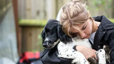 A black and white Spaniel dog being cuddled by a light-haired woman. The dog is looking to the right and the woman has her eyes closed and is snuzzling the dog's neck.