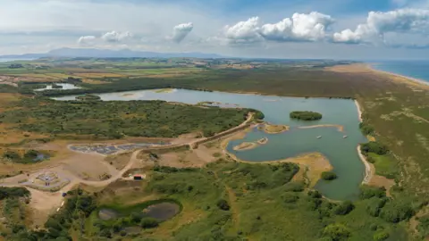 An ariel view of a nature reserve, there is a large pond in the middle, you can see the coastline on the other side of the reserve, on a clear blue day.