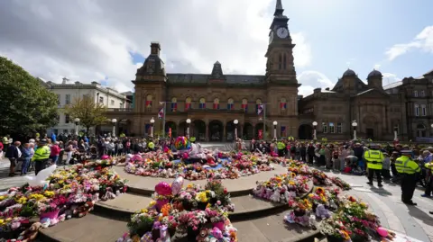 PA Media Floral tributes in a square outside The Atkinson arts centre in central Southport,  with large displays left on a circular stone area in the middle of a square in front of an ornate Victorian building in stone, with a clocktower at one end. Peaceful crowds are being marshalled by a handful of police officers 