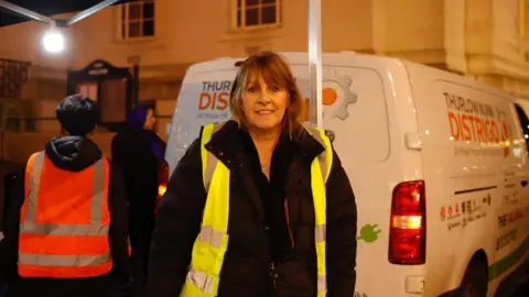 Kian Boyle/BBC A woman wearing a yellow hi-vis vest and dark coat stands under a lit canopy at night, facing the camera. Behind her, other volunteers in reflective vests work near a white van.
