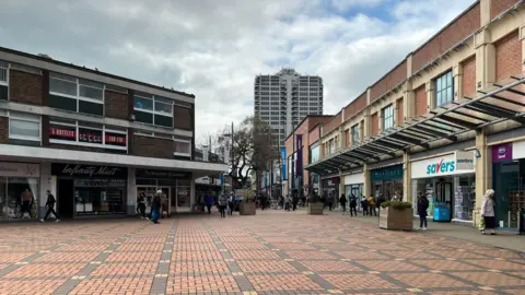 Swindon Town Centre on a grey day, tower block in the background, a pedestrianised area with shops on either side in red brick buildings two storeys high with flat roofs.