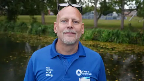A man standing by a river wearing a blue shirt with a Canal & River Trust logo, and sunglasses on his head. 
