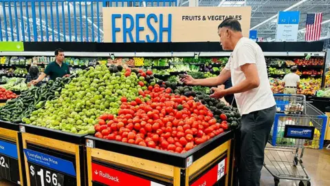 A man in a white T-shirt holds an avocado while standing in the produce section before a pile of tomatoes, avocadoes and other vegetables at a Walmart in Rosemead, California, on April 11, 2025.