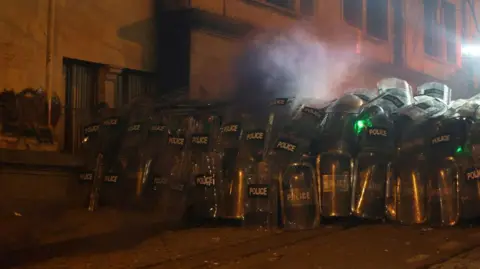 GIORGI ARJEVANIDZE/AFP Riot police wait on the street next to the parliament as tens of thousands of people protest in the distance