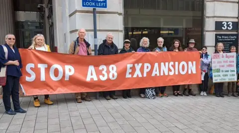 Adrian Howlett Group of campaigners stood holding an orange banner saying "Stop A38 Expansion" in Derby