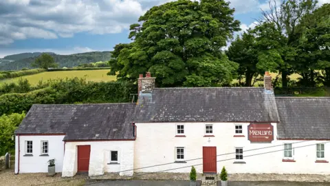 Under the Thatch Exterior view of the whitewash long cottage with two red wooden doors, small windows and slate roof, with a red pub sign, Maenllwyd