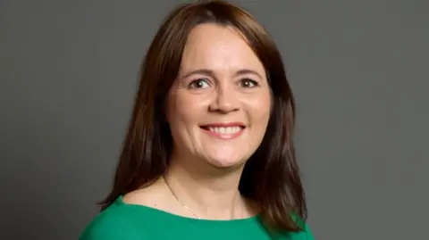 BBC Woman with long brown hair and brown eyes in green dress smiles in front of grey background