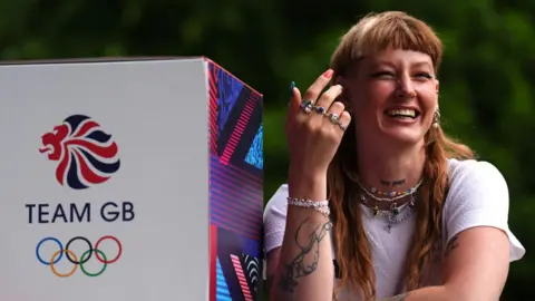 PA Media Makayla Gerken Schofield has long auburn hair and a fringe in this picture. She is wearing a white T-shirt and has a tattoo on their forearm. Next to them is a box that says "Team GB" with the Olympic rings on it.