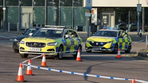 Police vehicles block off a section of road outside a modern glass-fronted building. Marked police cars are parked behind blue-and-white police tape and a line of orange traffic cones. Road signs, bollards and parked vehicles are visible around the cordon.