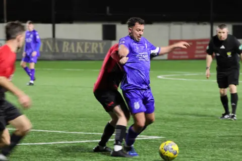 Claire Kirkpatrick A man in blue defends the ball from a player in red while the referee watches on