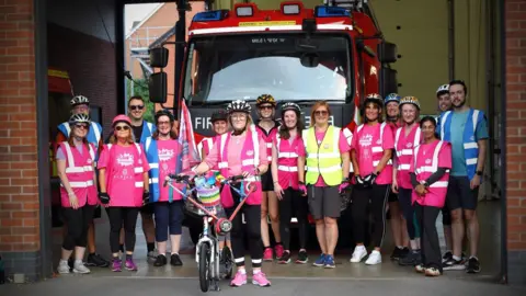 Supplied At the front of the photo is Rachel Liew on a scooter with a team of people stood behind her. They are at a fire station, and a fire engine is behind the group, who are all wearing either pink, blue or yellow hi vis jackets 