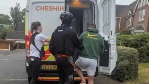 Two police officers escort a man with a green hooded top and white shorts into the back of a police van. The van is parked on a road with houses either side.