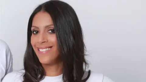 Nadine Buzzard-Quashie A woman with dark shoulder length hair stands in a white T-shirt. She has a broad smile. The background is white and indoors.