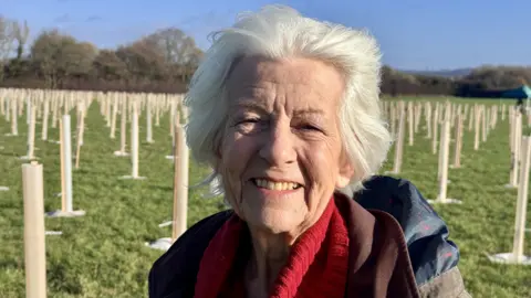 A woman with short white/grey hair is wearing a brown jacket and red jumper. She is smiling in the middle of a woodland where hundreds of trees are being planted.