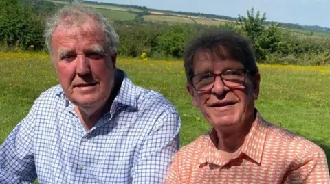 Martin Taylor Two men, one wearing a blue and white shirt, the other wearing a red and white shirt, are sitting together and smiling at the camera. Behind them is a field, a hedge and a countryside landscape in the distance.