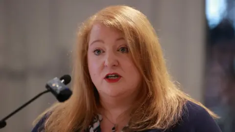 PA Media Justice Minister and Alliance person  Naomi Long has strawberry blonde and wearing a purple cardigan and speaking into a microphone.