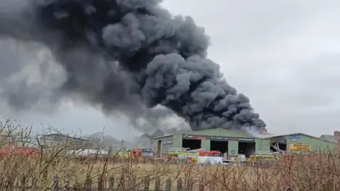 Stuart Peacock A tall, grey plume of smoke rises from a green metal industrial warehouse. There are fire engines parked out front. 