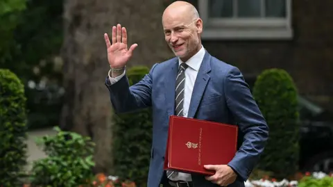 Stephen Kinnock arrives at Downing Street carrying a red Welsh secretary file and raising a hand in greeting. Trees, trimmed shrubs and flowerbeds can be seen in the background.