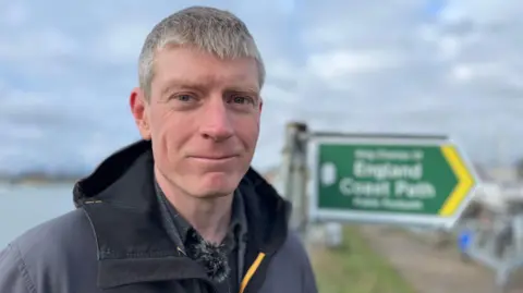 Graham Horton wearing a coat and has brown/grey hair and is standing on the path with a sign which reads "England coast path" behind him
