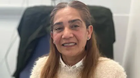 Nafisabanu Ahmed at the endoscopy centre at Leicester General Hospital. She has long brown hair and is looking at the camera. There is a chair behind her that has a black jacket.